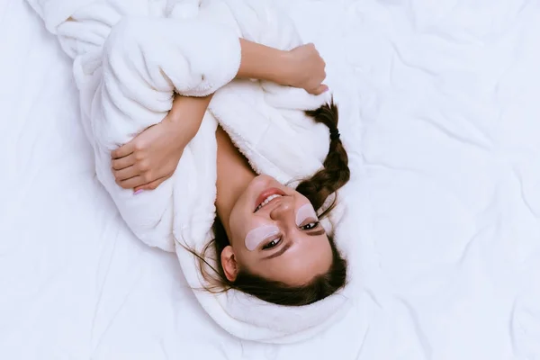 Uśmiechający się piękna dziewczyna w szlafrok biały frotte leży w łóżku wczesnym rankiem. pod oczami plastry Silikonowe — Zdjęcie stockowe