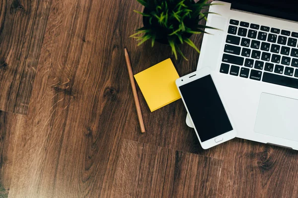 Деревянная поверхность - это ноутбук для работы, смартфон, желтая бумага и карандаш, зеленое растение — стоковое фото