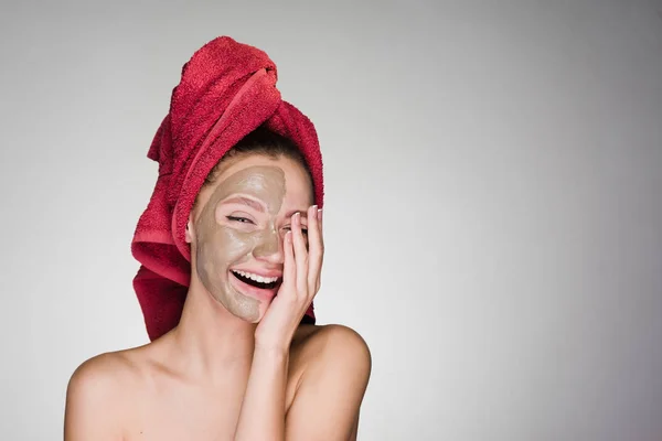 Skrattar vacker flicka med en röd handduk på hennes huvud, vita tänder, ansiktsmask hälften lera — Stockfoto