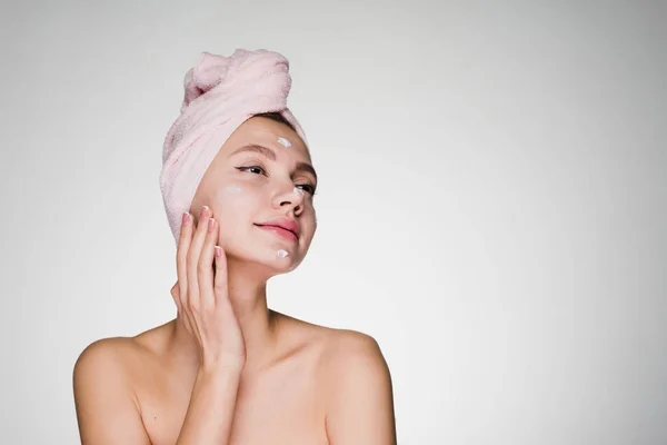 Jovem atraente com uma toalha na cabeça coloca no rosto um creme nutritivo branco — Fotografia de Stock