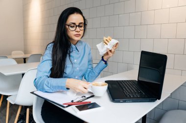 bir sandviç ve kahve ile bir kafede kahvaltı yiyor ve bir dizüstü bilgisayarda gözlük ile meşgul güzel kız serbest meslek