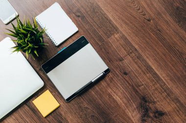 bir ahşap yüzeye bir dizüstü bilgisayar, bir grafik tablet, yeşil bir bitki, bir kağıt ve bir akıllı telefon var