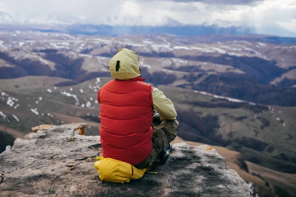 A male traveler in a jacket sits on the edge of a cliff and looks at the mountains, enjoys nature — Stock Photo, Image