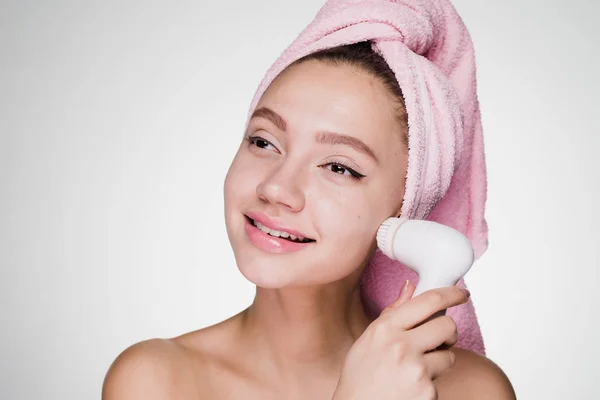 Menina sorridente feliz com toalha rosa na cabeça fazendo limpeza profunda da pele do rosto com escova elétrica especial — Fotografia de Stock