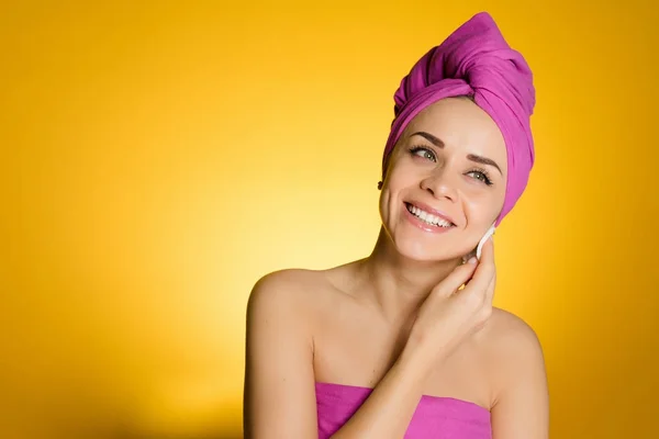 Ευτυχισμένος χαμογελαστό κορίτσι με μια ροζ πετσέτα στο κεφάλι της καθαρίζει το δέρμα του προσώπου της με ένα δίσκο βαμβακιού — Φωτογραφία Αρχείου