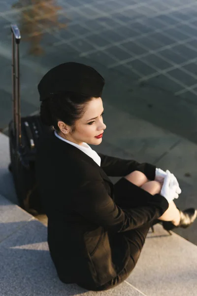 Promyšlené krásná letuška dívka v uniformě sedí na schodech v parku s kufrem a myslí na cesty — Stock fotografie