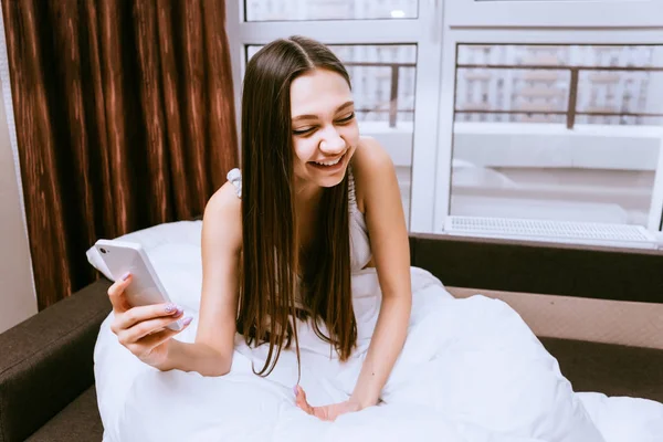 Смеющаяся длинноволосая девушка, сидящая в постели под одеялом, держа в руке смартфон — стоковое фото