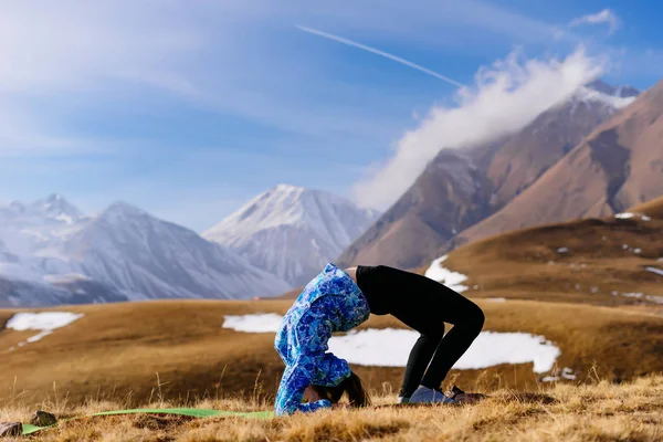 Гибкая молодая девушка в синей куртке занимается йогой на фоне Кавказских гор — стоковое фото