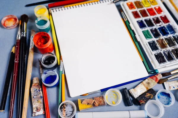 On a blue table are artistic accessories, watercolors and gouache, many brushes and white paper — Stock Photo, Image
