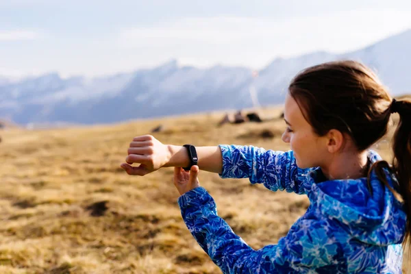 Mladá žena cestovatele v modrém saku vypadá v době na hodinky na pozadí kavkazských hor — Stock fotografie