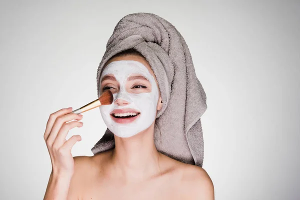 Hermosa chica sonriente pone una máscara blanca nutritiva en su cara, en su cabeza una toalla —  Fotos de Stock
