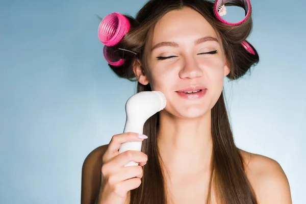 Uma jovem satisfeita faz uma limpeza profunda da pele em seu rosto com uma escova elétrica, em seus encrespadores de cabeça — Fotografia de Stock