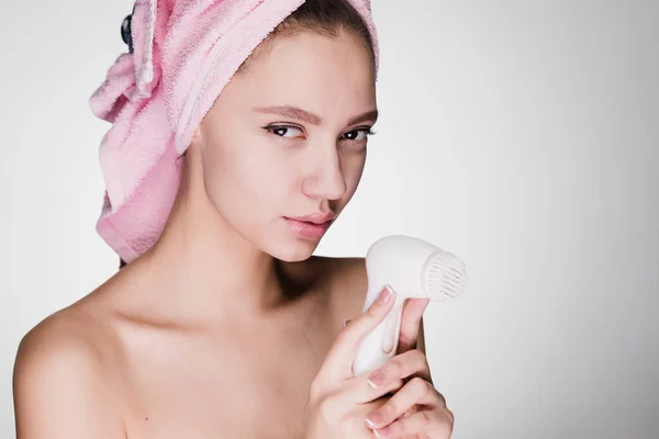 Attractive young girl with a pink towel on her head holds a special electric brush for deep cleansing of the skin on her face — Stock Photo, Image