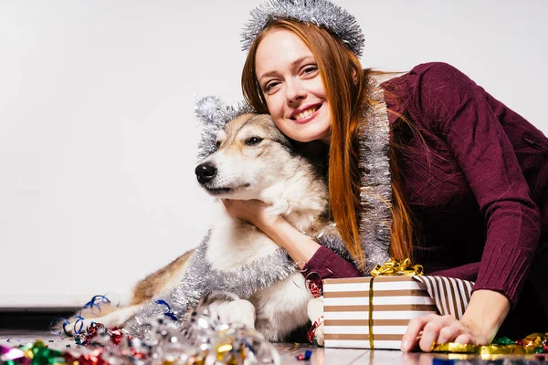 Vermelho de cabelos sorridentes menina abraçando seu cão para o ano novo 2018, ouropel de prata e presentes — Fotografia de Stock