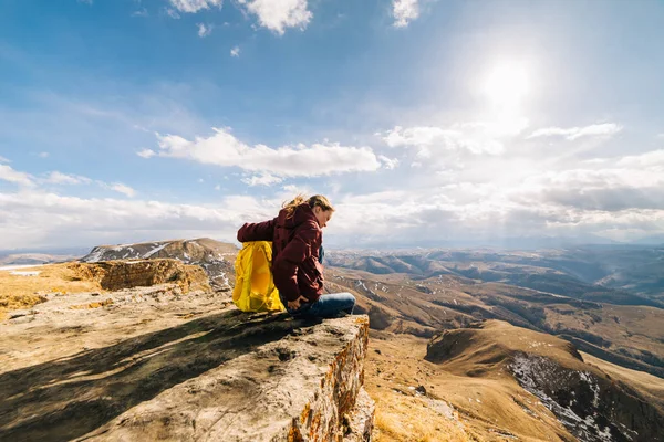 Una joven con una mochila amarilla se sienta en el borde de un acantilado. disfruta de la naturaleza y el aire limpio de montaña —  Fotos de Stock