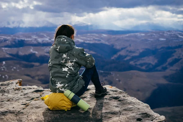 Молода дівчина в теплій куртці сидить на краю гори, насолоджуючись природою і чистим повітрям, чаєм в термосі — стокове фото