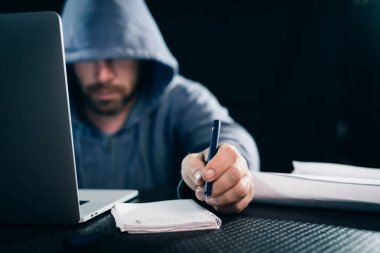 gizemli tehlikeli erkek hacker yüzünü başlık altında gizler, bir dizüstü yanında bir şey yazıyor