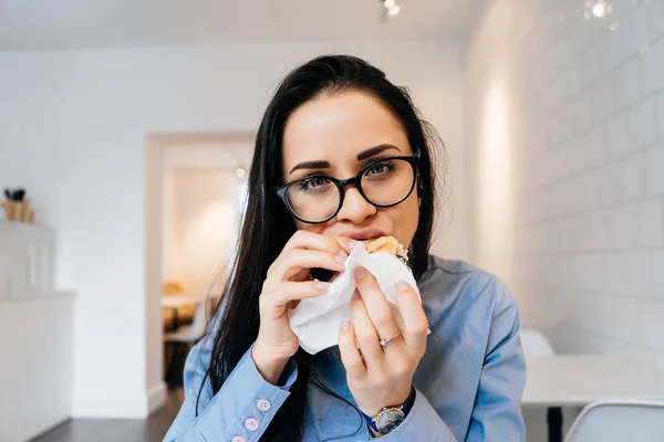 Молодая голодная девушка в очках охотно ест сэндвич в кафе после работы. — стоковое фото