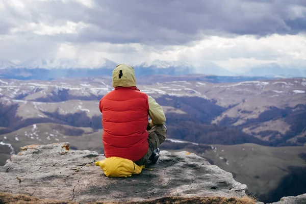 Viajero masculino en una chaqueta roja se relaja, se sienta en el borde de un acantilado y mira a las montañas —  Fotos de Stock