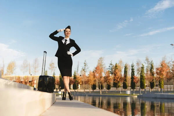 Улыбающаяся молодая стюардесса салютует, с чемоданом, в парке — стоковое фото