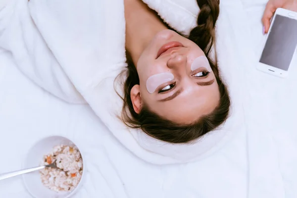 Joven hermosa chica en una túnica blanca se encuentra en una cama temprano en la mañana, bajo los ojos de un parche, junto a un plato con cereales para el desayuno —  Fotos de Stock