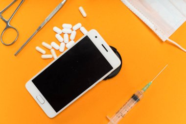 Turuncu yüzeyinde beyaz tabletler, bir akıllı telefon, bir şırınga ve diş aletleri vardır