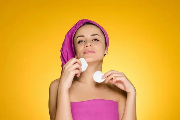 Attractive young girl with a pink towel on her head cleans the skin on her face with cotton pads — Stock Photo, Image