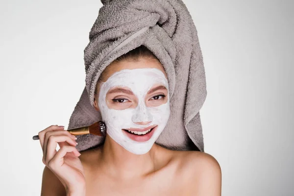 Beautiful young girl with a towel on her head puts a white mask on her face with a brush — Stock Photo, Image