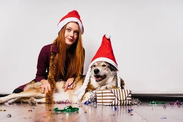 Red-haired 漂亮的女孩, 戴着红帽子, 脖子上戴着金色的金属箔, 和她的狗一起坐在地板上, 等待新的一年 — 图库照片