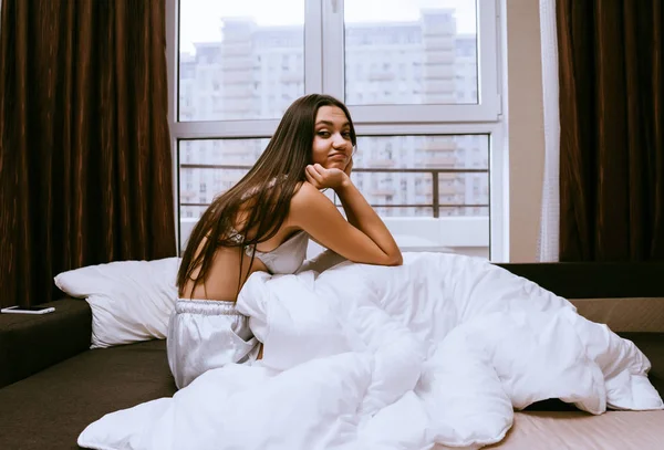 Привлекательная молодая девушка просыпается рано, сидит в постели под белым одеялом — стоковое фото