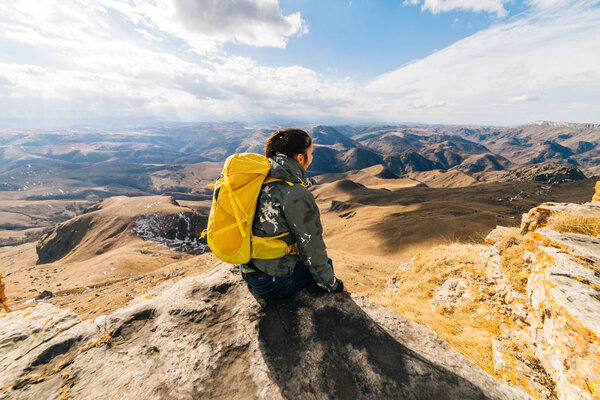 A young active girl sits on the edge of the cliff, admires the mountains and enjoys pure air