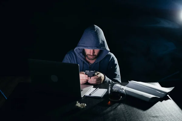 Mystérieux homme criminel dans un pirate de capot, cache son visage, près d'un ordinateur portable, écrit quelque chose sur smarfone — Photo