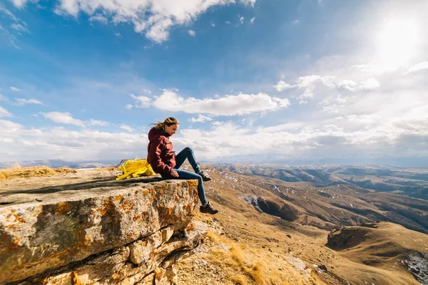 Una chica joven lleva una forma de vida activa y saludable, viaja, se sienta en un acantilado y disfruta de la naturaleza de montaña —  Fotos de Stock