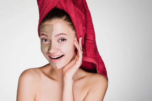 Feliz hermosa chica con una toalla roja en la cabeza se aplica una máscara de arcilla a la mitad de la cara — Foto de Stock