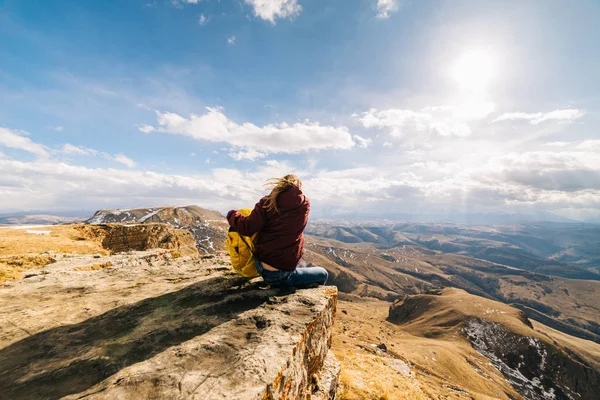 Молодая девушка сидит на краю горы, путешествует по Кавказскому хребту с рюкзаком — стоковое фото