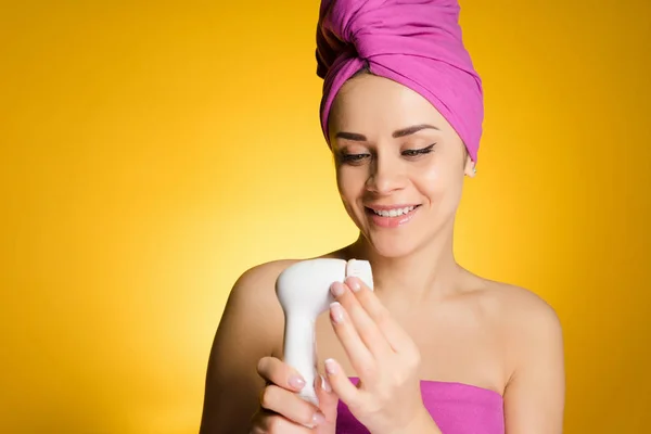 Χαμογελώντας όμορφο κορίτσι με μια ροζ πετσέτα στο κεφάλι της, σε κίτρινο φόντο, κρατώντας ένα ηλεκτρικό Βουρτσάκι για βαθύ καθαρισμό κωδικοποίησης στο πρόσωπό της — Φωτογραφία Αρχείου