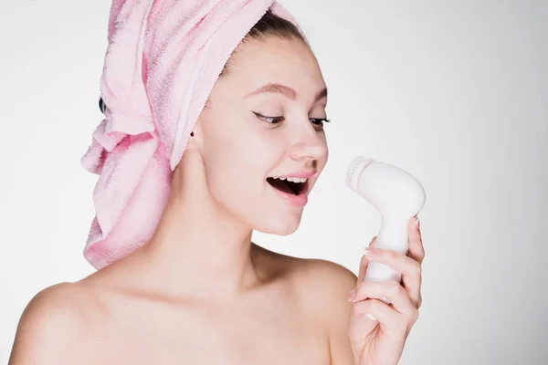 Una ragazza sorridente si prende cura del suo viso con un asciugamano rosa sulla testa, tiene in mano una spazzola elettrica per una pulizia profonda del codice sul suo viso — Foto Stock