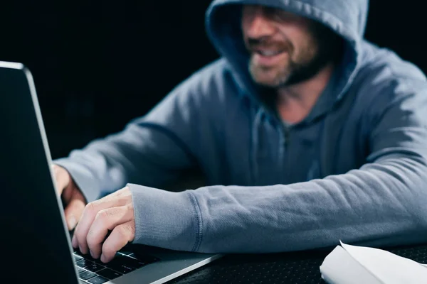 the grinning mysterious man hides his face under the hood, hacks something on the laptop