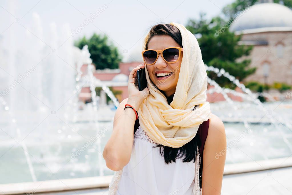 A young tourist girl in a headscarf on her head uses a phone, stares into the distance in sunglasses