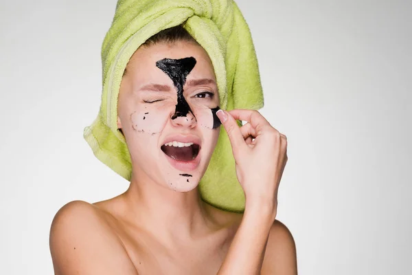 Молода дівчина з зеленим рушником на голові видаляє чорну маску для очищення обличчя, їй незручно — стокове фото