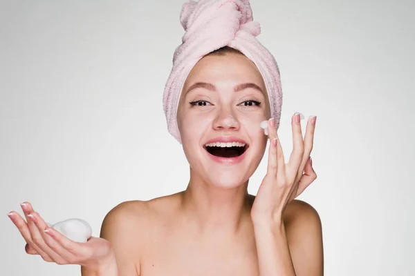 Menina feliz com uma toalha rosa na cabeça e dentes brancos aplicar espuma de limpeza em seu rosto — Fotografia de Stock