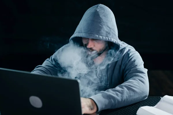 Μυστηριώδης άνδρας χάκερ κρύβει το πρόσωπό του κάτω από την κουκούλα, καπνίζει, ο πολύς καπνός — Φωτογραφία Αρχείου