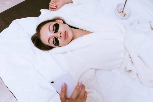 Uma menina em um manto branco jaz na cama, acordou cedo, sob os olhos de manchas pretas, faz selfie — Fotografia de Stock