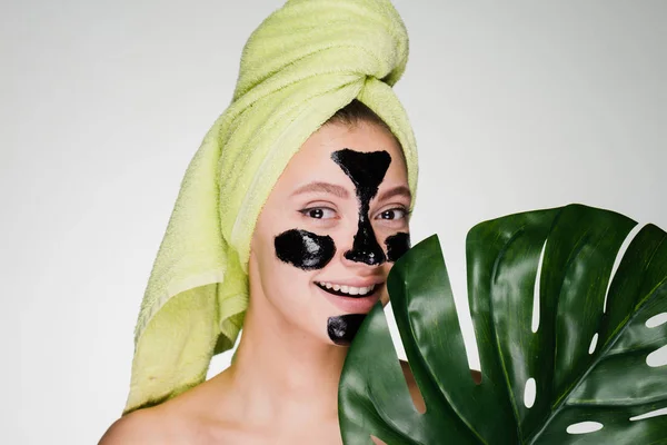 A smiling beautiful girl with a green towel on her head applied a black cleansing mask to the problem areas on her face — Stock Photo, Image