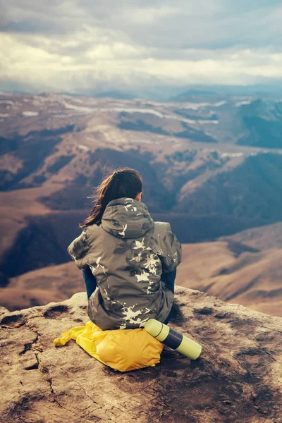 Chica se sienta en el borde de la montaña en una mochila amarilla, disfruta de la naturaleza y el aire de la montaña, té caliente en un termo —  Fotos de Stock