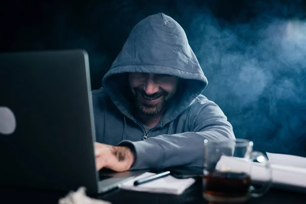 a grinning criminal man hides his face under the hood and hacks something on the laptop