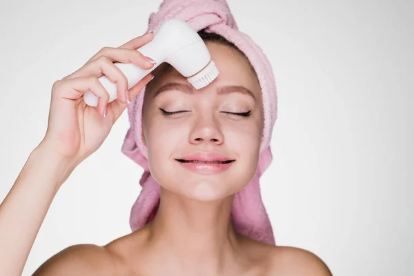 Smiling happy woman with a pink towel on her head cleans the skin on her face with a special electric brush — Stock Photo, Image