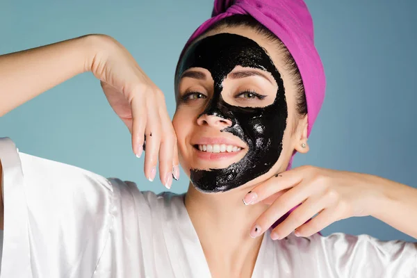 Bella ragazza sorridente con un asciugamano rosa sulla testa vuole una pelle bella e pulita, ha applicato una maschera di pulizia nera sul suo viso — Foto Stock