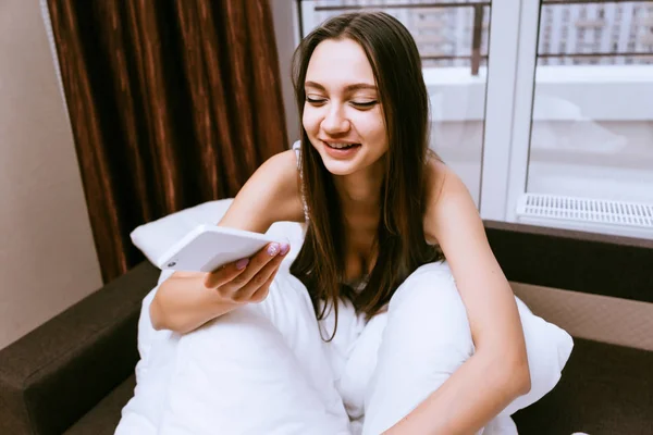 Menina com um telefone em mãos sorrindo para o fundo da janela — Fotografia de Stock