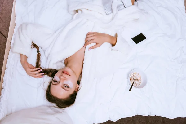 En flicka i en mantel ligger på en säng med en mask under ögonen — Stockfoto